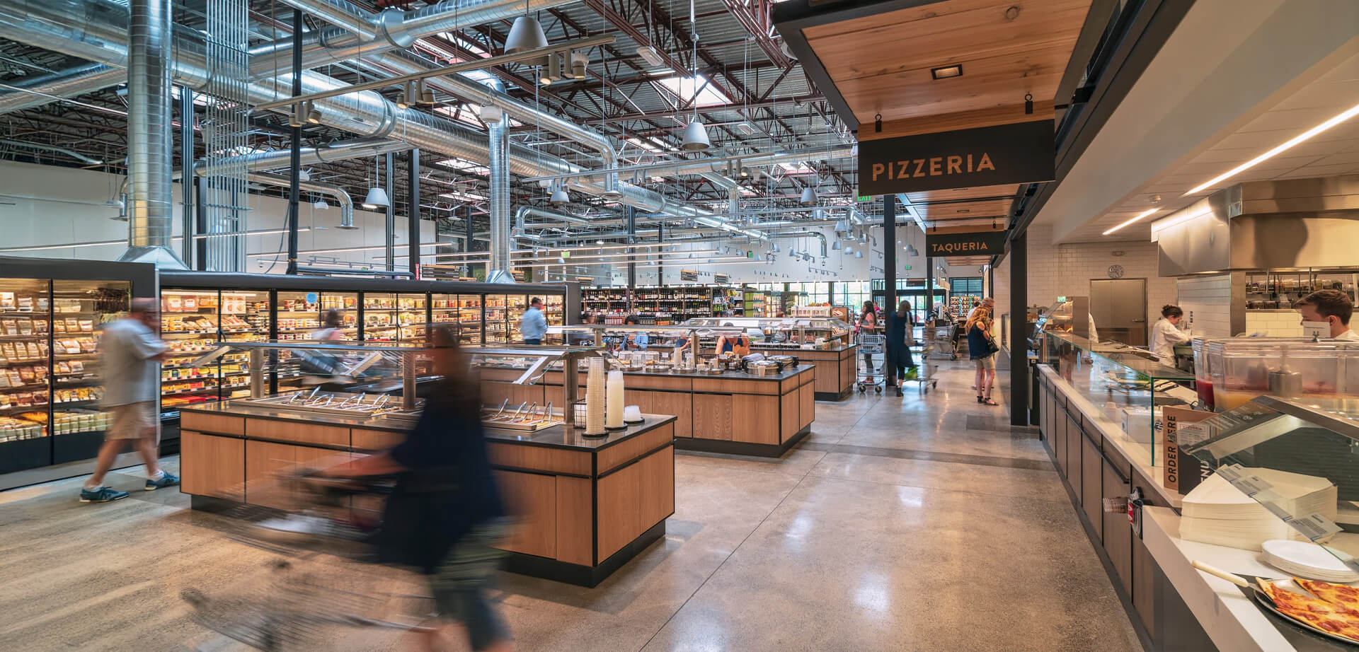 The image is an interior view of a PCC Community Markets grocery store. These stores are known for their sustainable building practices and focus on local products.  The store features an open industrial-style ceiling with exposed pipes and ductwork.  There are dedicated sections for prepared foods, including a Pizzeria and Taqueria.  PCC Community Markets is a community-owned food market chain primarily located in the Seattle, Washington area. 