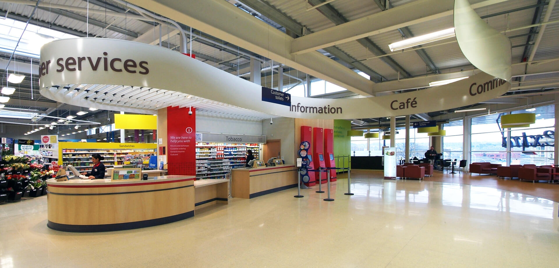 The Tesco Extra Hypermarket Accrington, Lancashire England. Clear Signage and Wayfinding: Prominent, clearly labeled department signs (like the "Customer Services", "Information", and "Café" signs in the image) help customers navigate easily and reduce anxiety. This ensures shoppers can find specific items quickly.