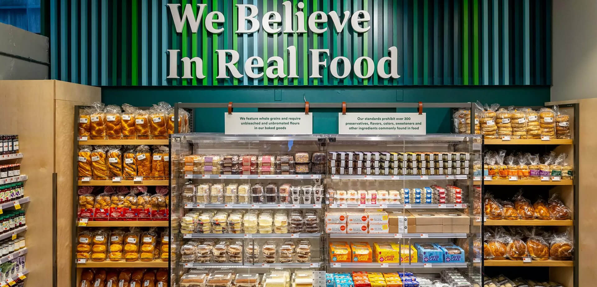 The baked goods section at a Whole Foods Market, highlighting the store's "We Believe In Real Food" philosophy and quality standards. The text emphasizes the use of specific ingredients and the avoidance of common artificial additives. 