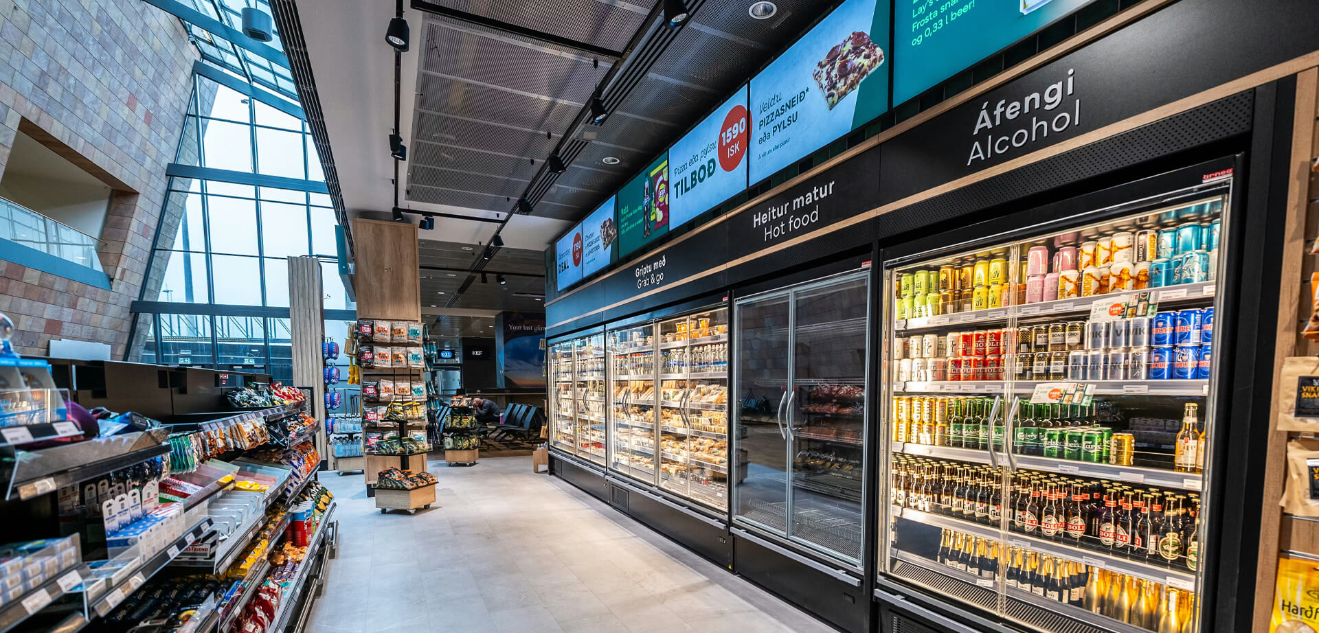 This image appears to be the interior of a Point KEF Airport store in Iceland. This convenience store offers a variety of travel essentials, snacks, and beverages, including both "Heitur matur" (hot food) and "Áfengi" (alcohol), as indicated by the Icelandic signage.