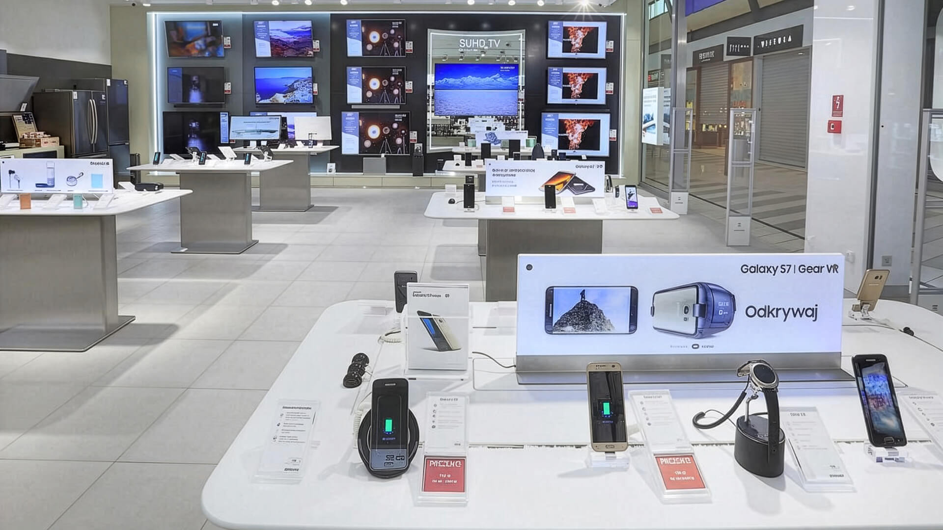 The image shows a modern Samsung Experience Store displaying various smart devices, including the Samsung Galaxy S7, Gear VR headsets, and a variety of SUHD TVs on the wall.