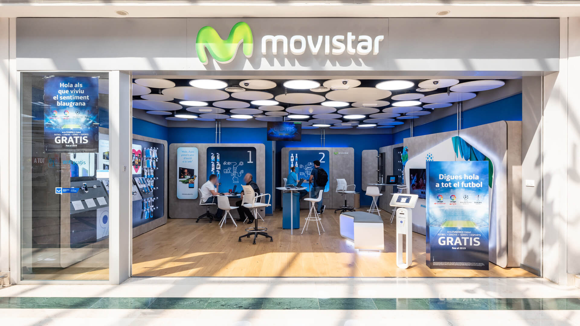This image shows a modern Movistar telecommunications store inside a shopping mall, featuring an open glass storefront, illuminated circular ceiling panels, and promotional displays for mobile and television services