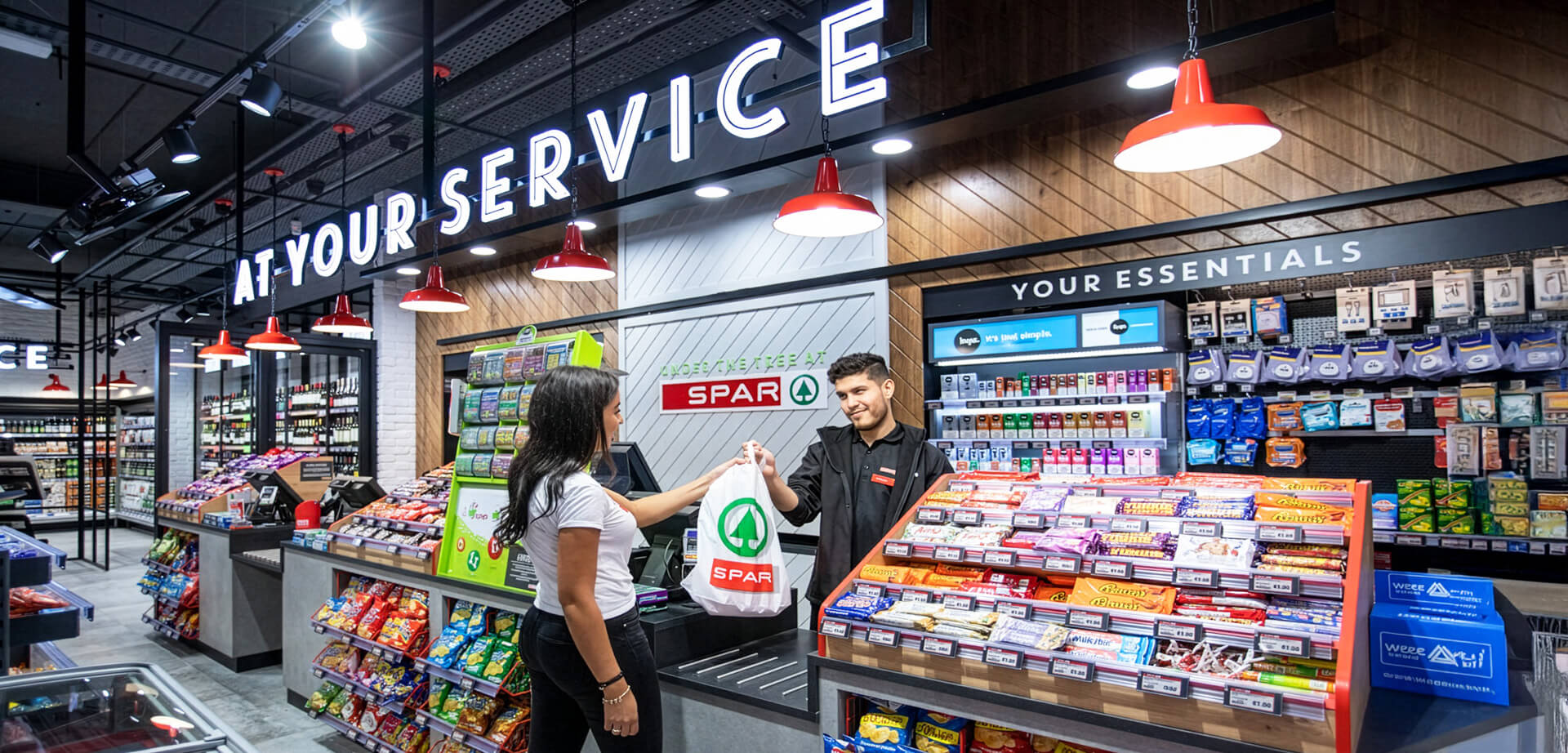 This image showcases a modern SPAR Ireland convenience store, highlighting the vibrant checkout area and the "Your Essentials" section stocked with everyday goods. The store is designed with sleek wooden panels, polished floors, and bright lighting to create a welcoming shopping environment.