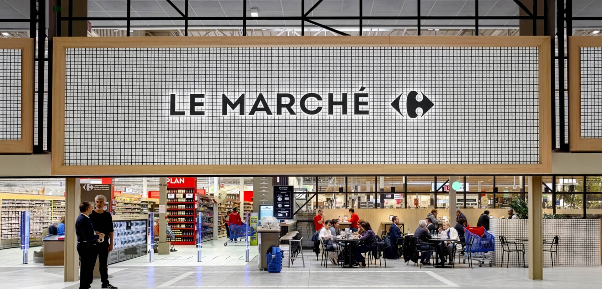 This image shows a redesigned fresh food and dining section inside a Carrefour hypermarket, featuring the "Le Marché" sign, wooden paneling, a grid-patterned backdrop, and an open seating area where customers can eat and socialise.
