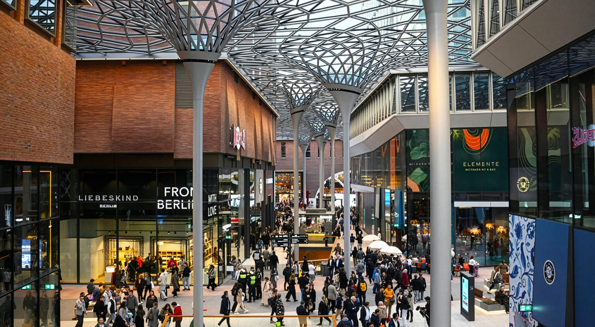 This image shows the bright, open pedestrian arcade of the Westfield Hamburg-Überseequartier lifestyle and shopping destination in Hamburg's HafenCity. Architecture: The space features a distinctive, tree-like column and geometric canopy roof that shelters the open-air style walkway while letting in daylight.