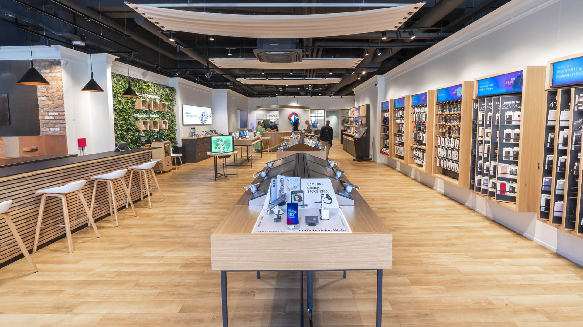 Swisscom Store layout: Open-plan design with light wooden flooring and central wooden display tables.Displays: Wall-mounted shelves showcasing mobile phones and tech accessories on the right side.Seating Area: A service or tasting counter with white bar stools on the left.Decor: A vertical green plant wall and black pendant lamps hanging from the ceiling.