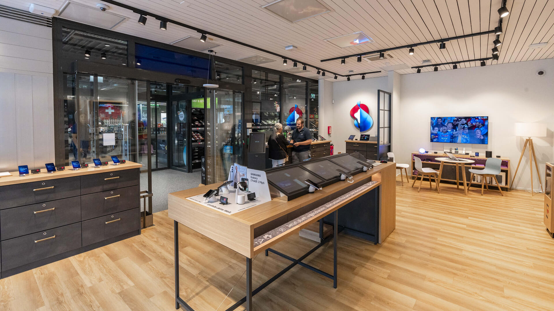 The image shows the interior of a retail telecommunications store, a Swisscom Shop, featuring dedicated brand display sections for Apple and Samsung with interactive product tables and wall fixtures.