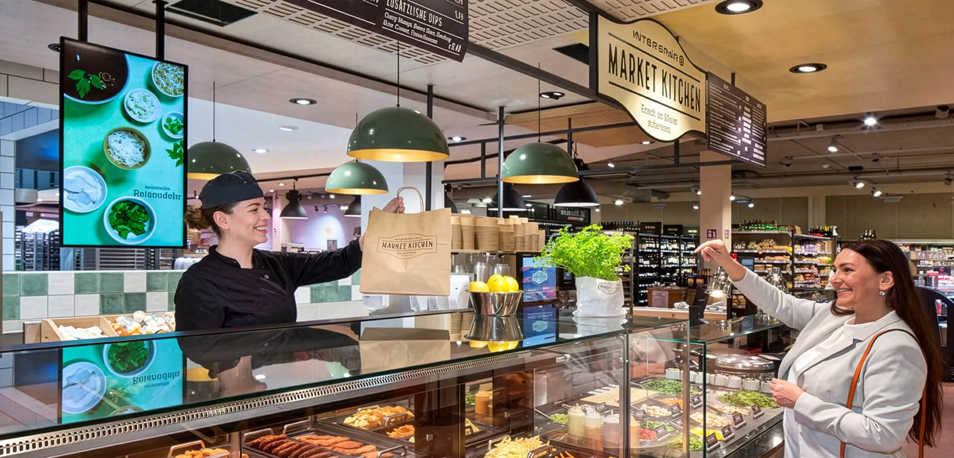 This image shows an Interspar Market Kitchen food counter where a staff member hands a paper bag to a customer.